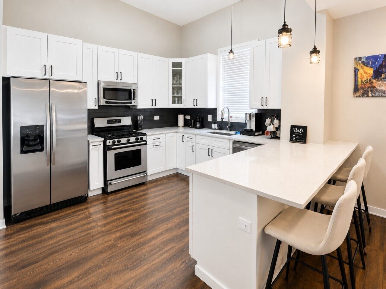 A modern kitchen with white cabinets, stainless steel appliances, a central island with bar stools, and pendant lighting.