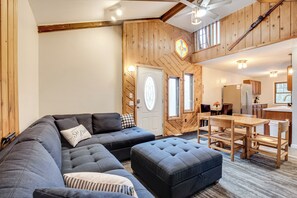 Een moderne woonkamer met een hoekbank, een voetenbankje, houten details, en een eethoek met een tafel en stoelen.