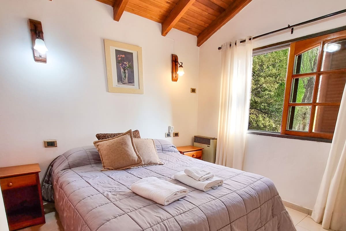 A bedroom with a large bed, a nightstand, a bedside table, a wall-mounted light, a framed picture, and a window with curtains.