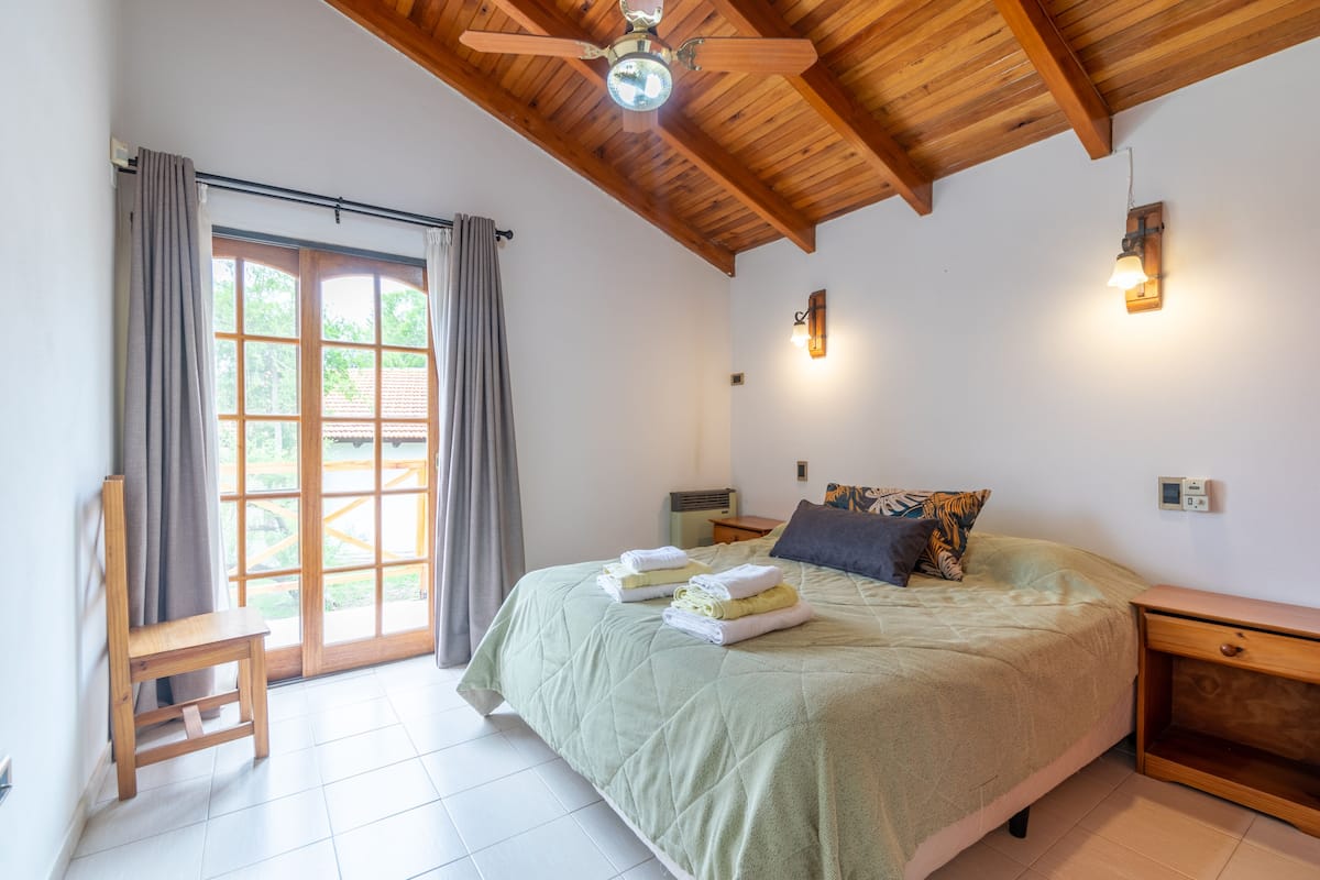 A bedroom with a bed, a wooden ceiling fan, a nightstand, and a window with curtains.