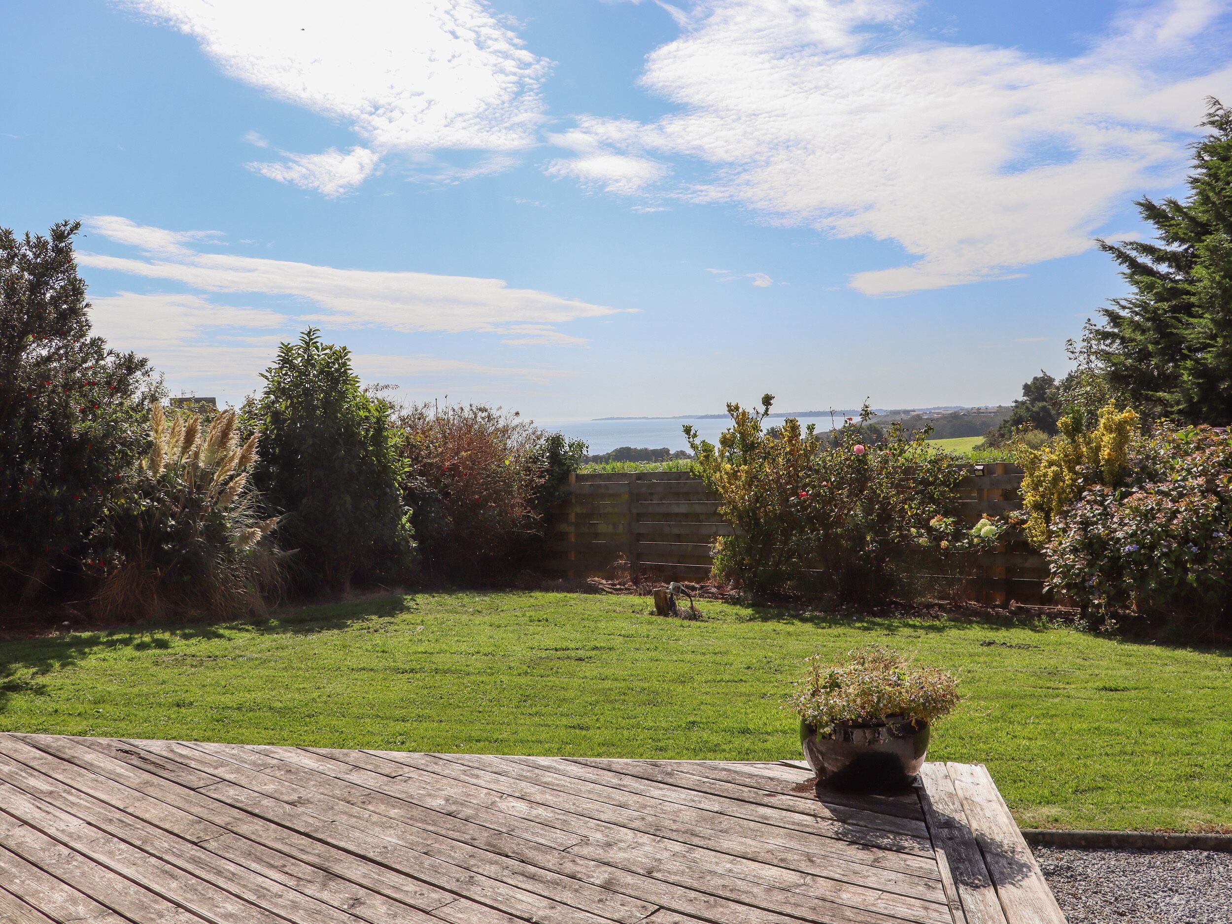 Una terraza de madera con una planta en maceta, un césped y vistas a un cuerpo de agua y colinas al fondo.
