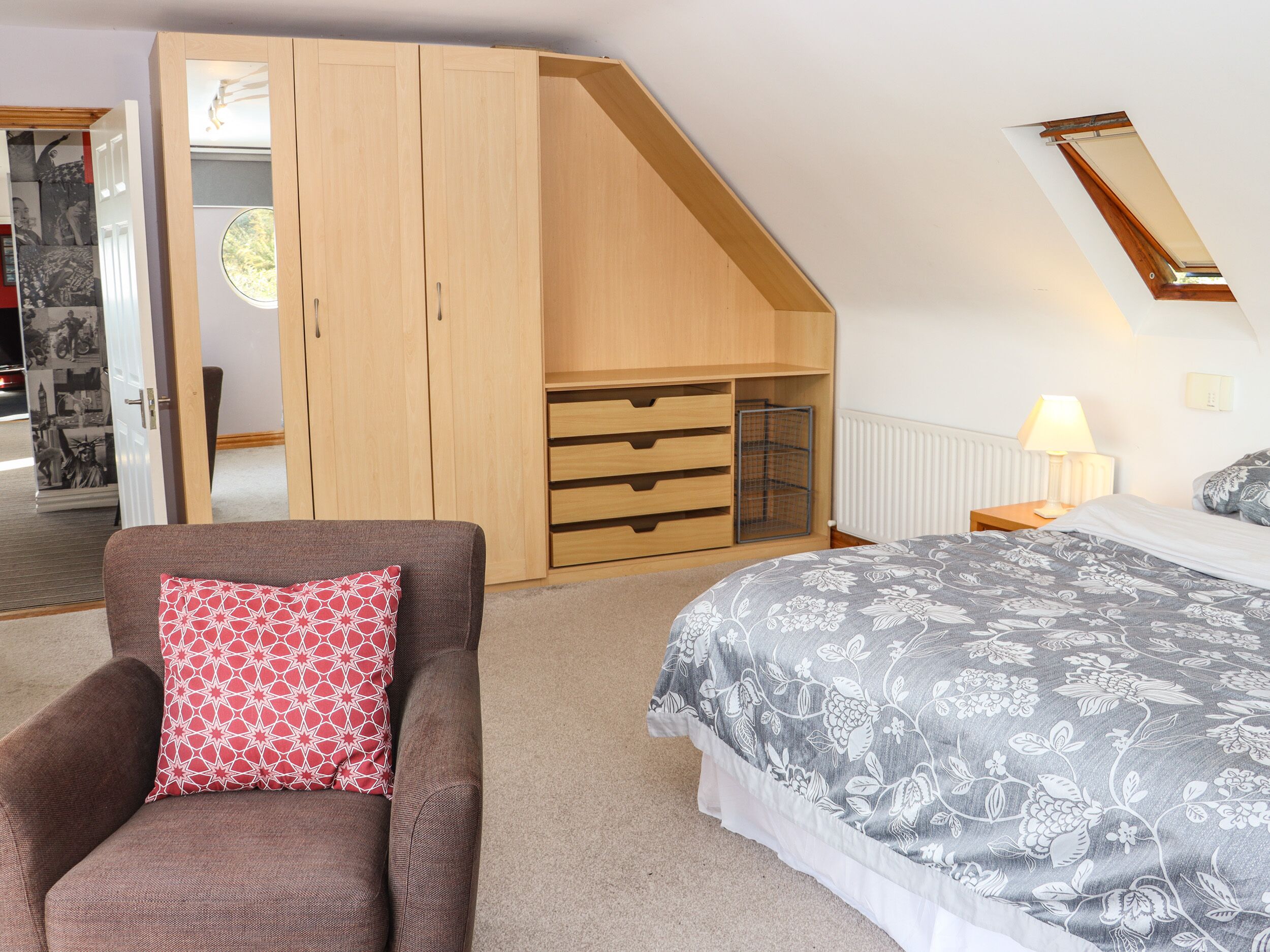 Un dormitorio con techo inclinado, una cama con colcha estampada, un sillón marrón con cojín rojo, un armario de madera y un ventanal pequeño.