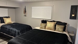 A bedroom with two beds, each with black and gold bedding, a window with blinds, a small table, and a framed picture on the wall. - The Superior Lodge - Located On Trail - Pictured Rocks Lakeshore, Munising MI (Munising)