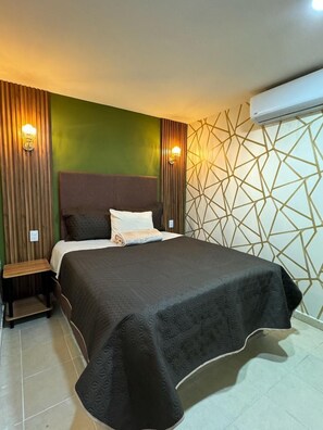 Uma cama bem arrumada com cobertura escura, uma mesa de cabeceira e duas luminárias fixadas na parede.