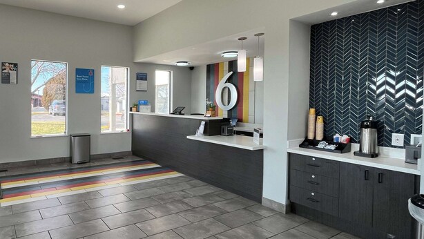 A modern reception area with a counter, a large number '6' on the wall, and a herringbone-patterned wall behind it. - Studio 6 Ogden, Ut – 21St Street (Ogden)