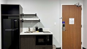A compact kitchenette with a black refrigerator, a sink, and a microwave. There's a wooden door with a fire evacuation plan sign, and a trash bin is visible. - Studio 6 Ogden, Ut – 21St Street (Ogden)