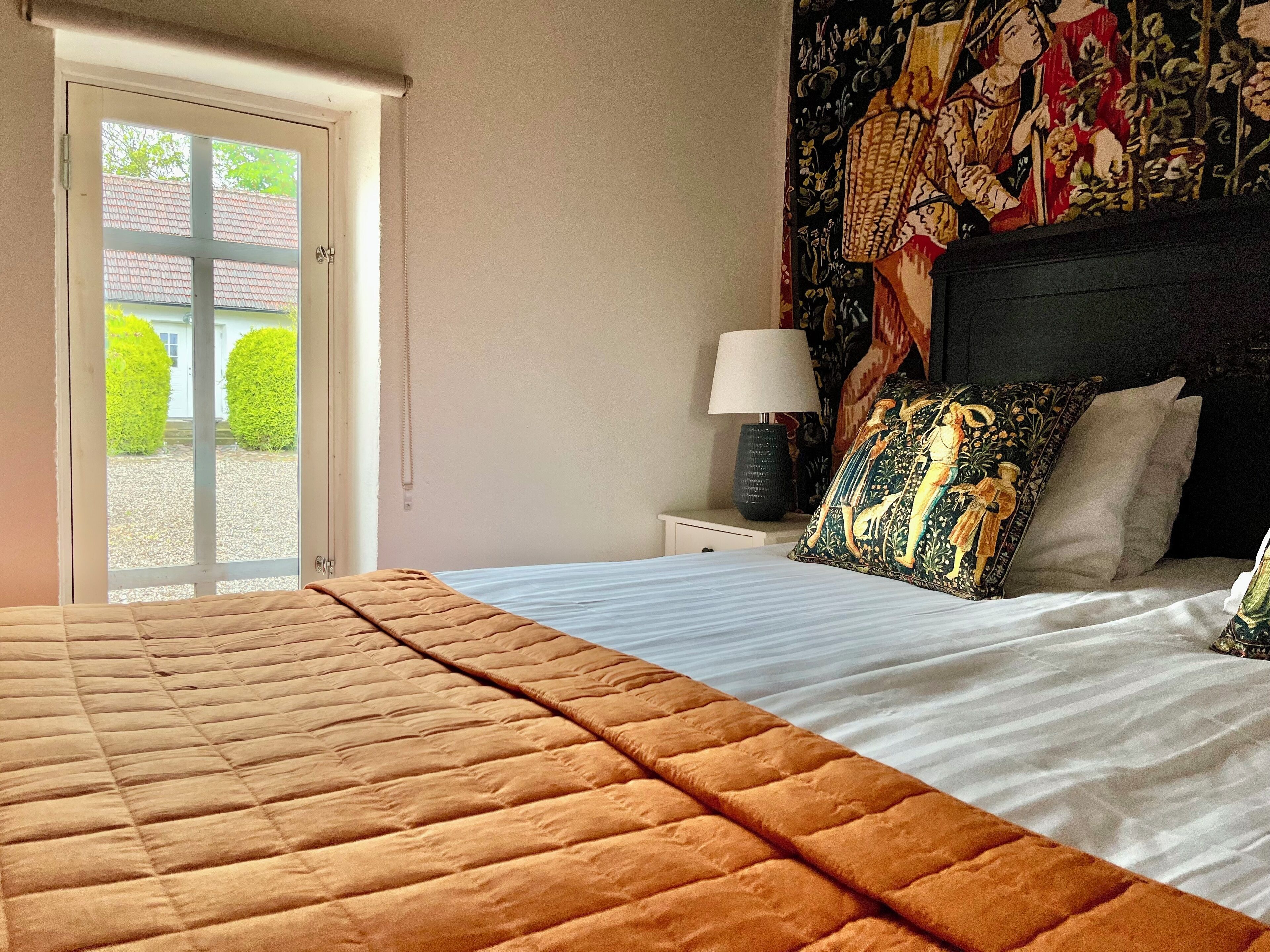 Un dormitorio con cama, mesita de noche con lámpara, ventana con vista a zonas verdes y un tapiz decorativo en la pared.