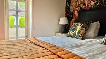 Un dormitorio con cama, mesita de noche con lámpara, ventana con vista a zonas verdes y un tapiz decorativo en la pared.
