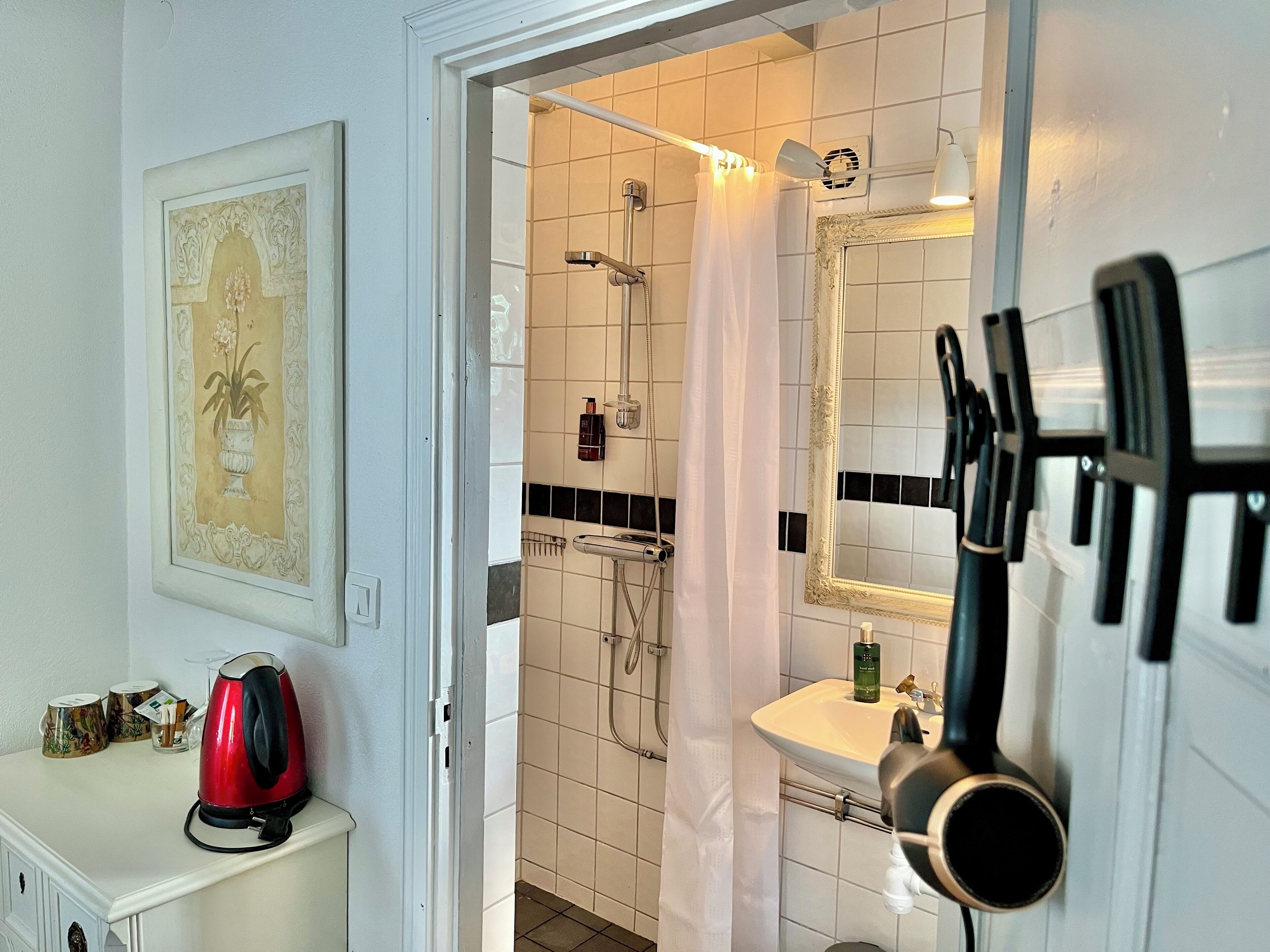 Une salle de bain avec une douche, un lavabo et un miroir. Un tableau encadré est accroché au mur et une bouilloire rouge est posée sur le comptoir.