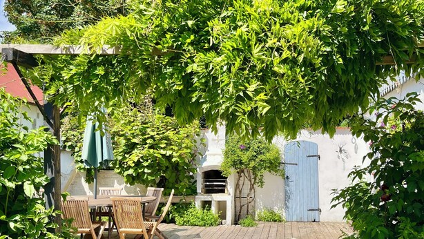 A wooden deck with a table and chairs, surrounded by lush greenery and a blue door.