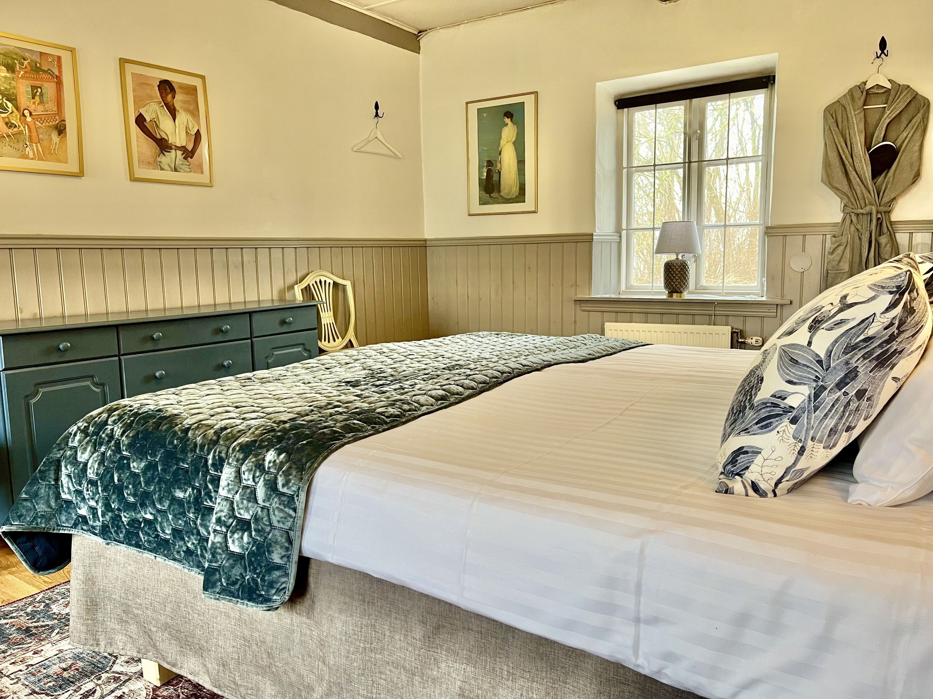 A bedroom with a bed, dresser, window, and artwork on the wall.