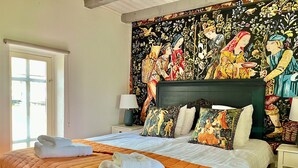 A neatly made bed with an orange quilt, white pillows, and folded towels. A large tapestry with a detailed scene hangs above the bed.