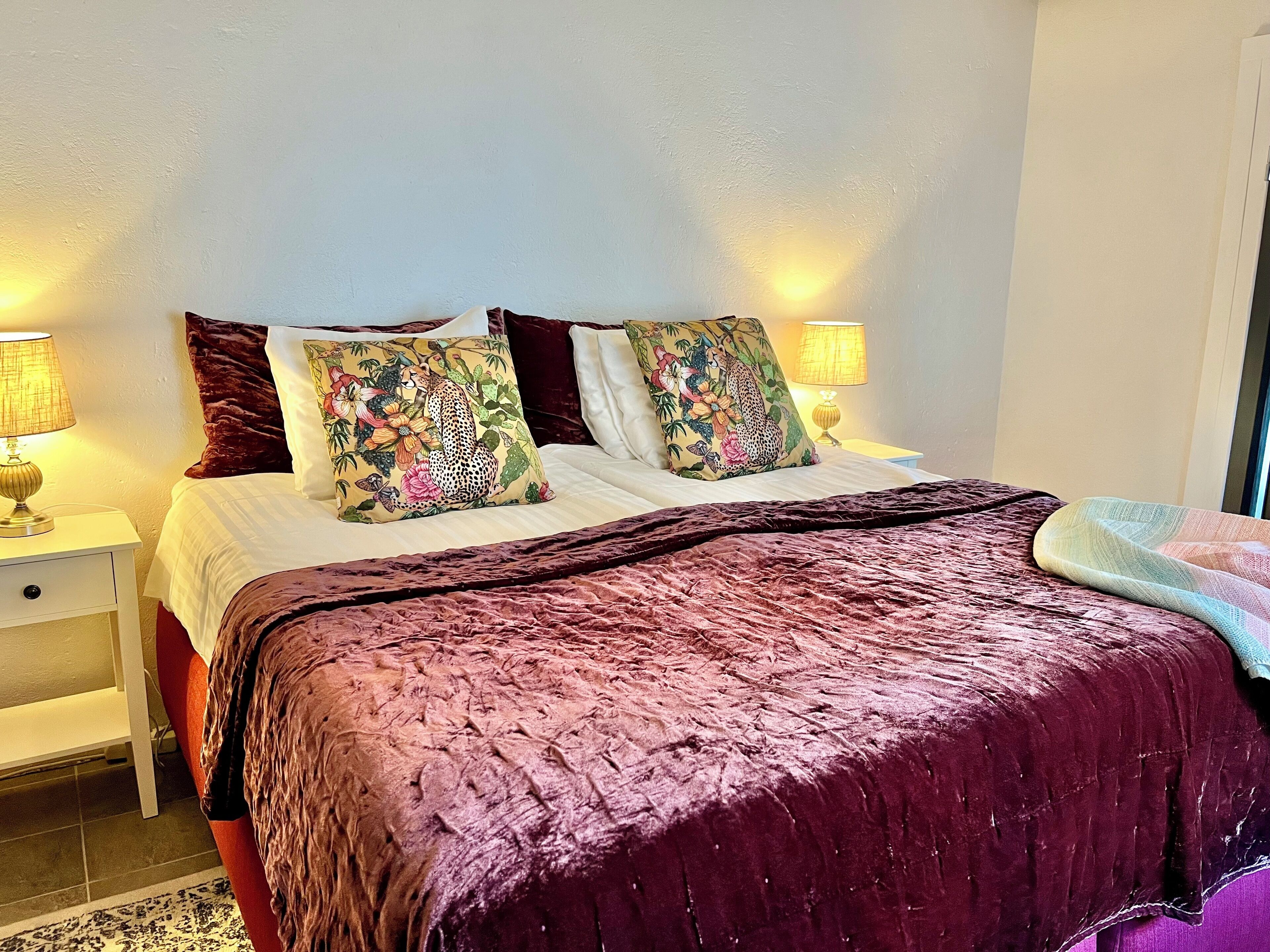 Una cama con un edredón morado, dos mesitas de noche con lámparas y almohadas estampadas con motivos florales.