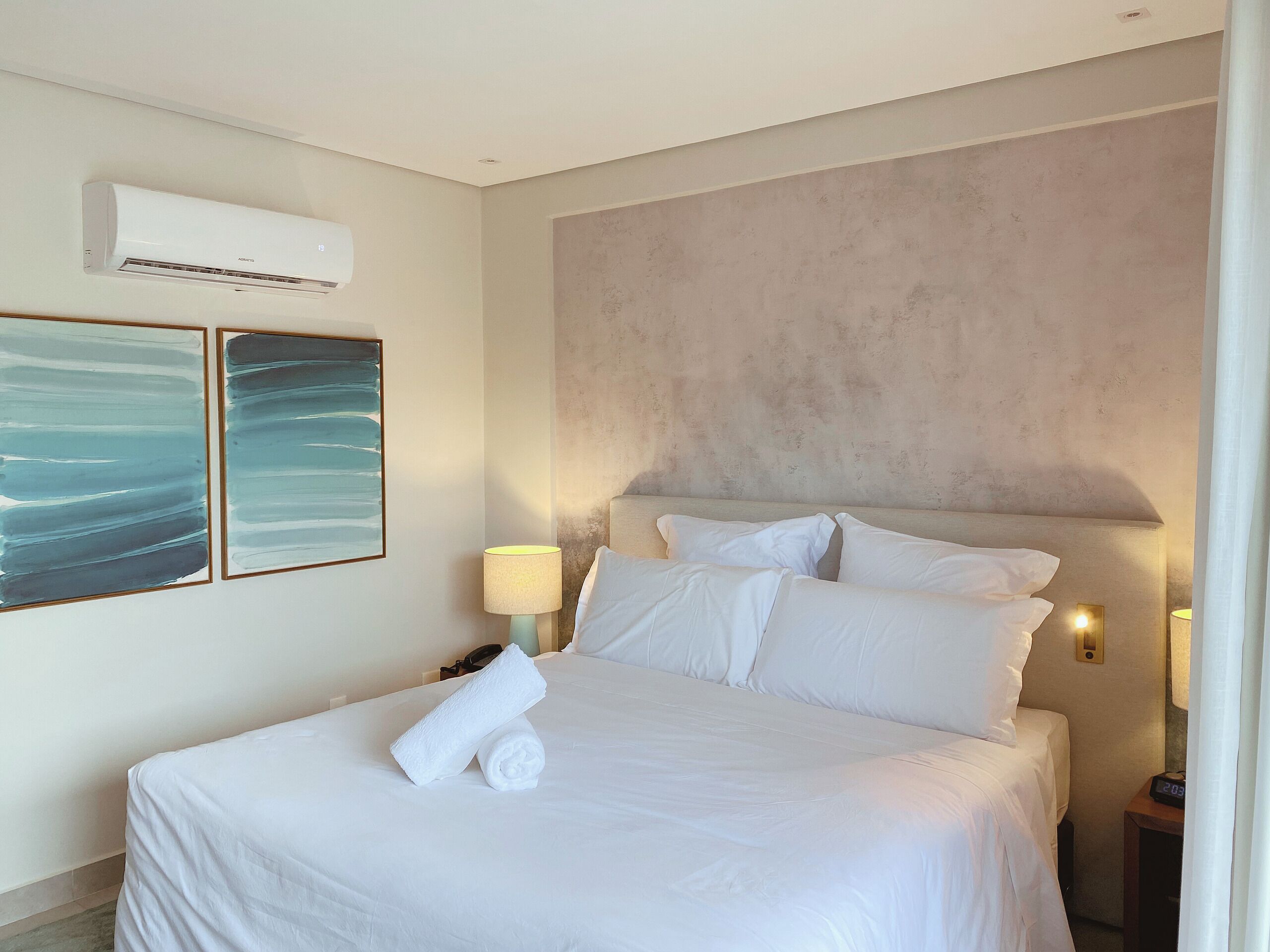 Ein Hotelzimmer mit einem Bett, weißen Leinen, zwei abstrakten Gemälden, einer Klimaanlage und einem Nachttisch mit einer Lampe.