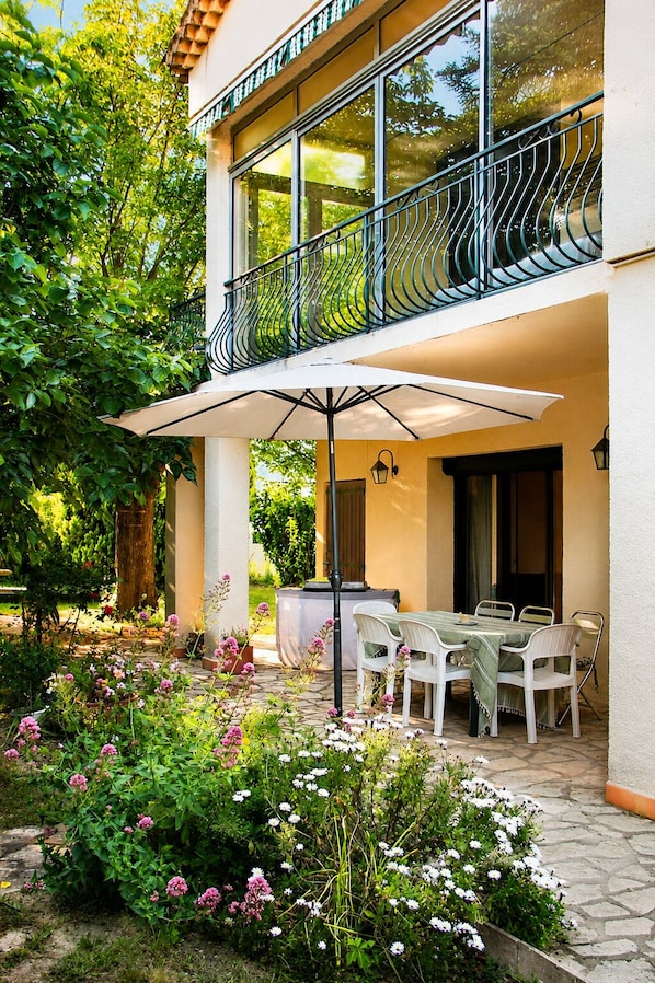 A two-story building with a balcony, outdoor dining area with chairs and a table, and a garden with various plants and flowers.