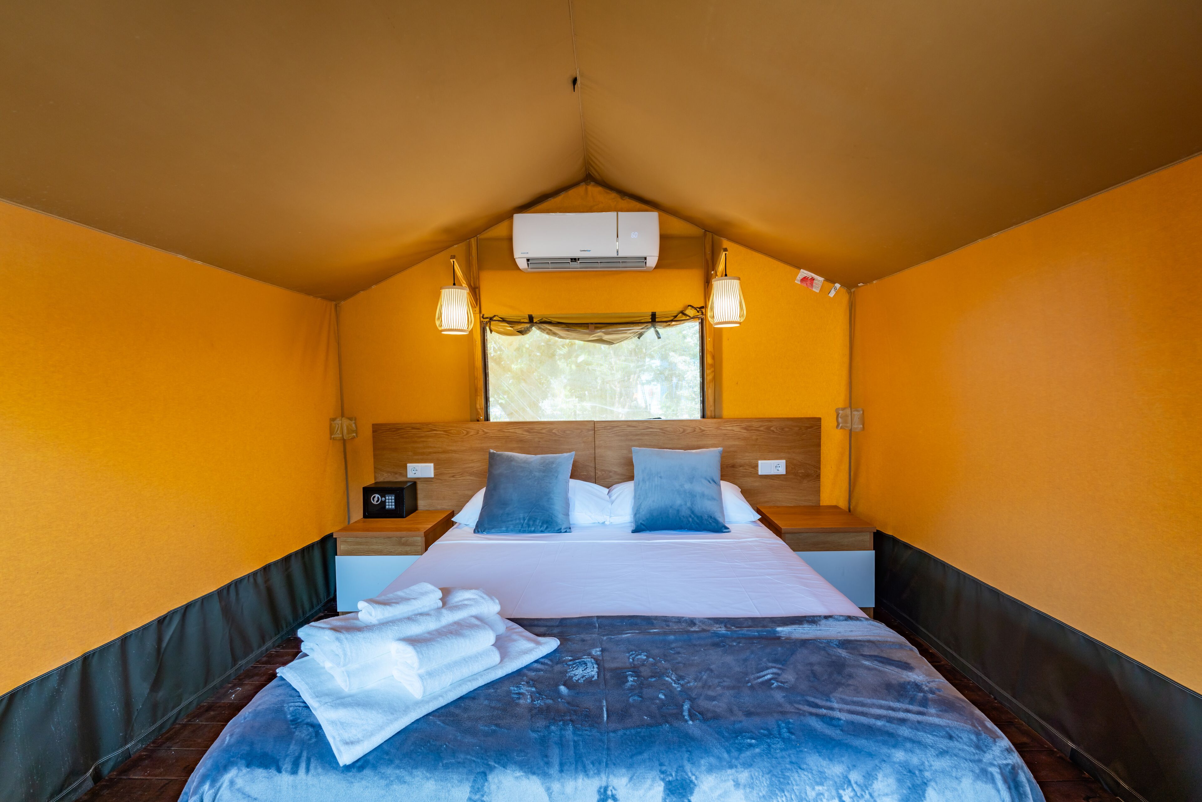 A single bed with a headboard, two bedside tables, an air conditioning unit, and a window with curtains.