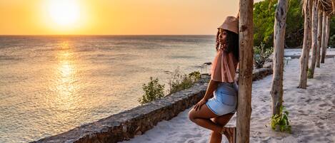 A beach scene with a sunset, a person standing near a wooden structure, and a stone wall.