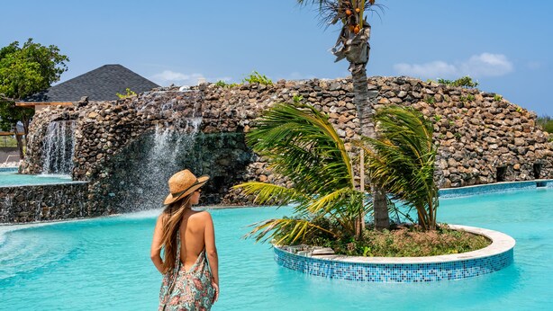 Mulher de vestido estampado e chapéu de palha parada perto de uma piscina com cascata e palmeiras.