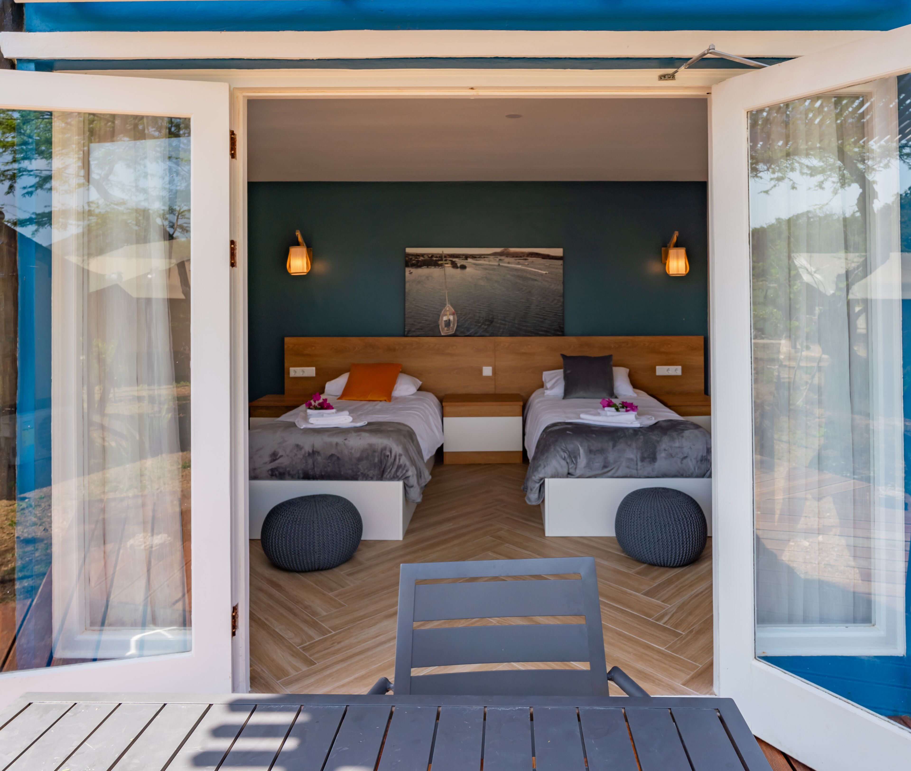A modern bedroom with two beds, a wooden headboard, a painting, and a view of the outdoors.