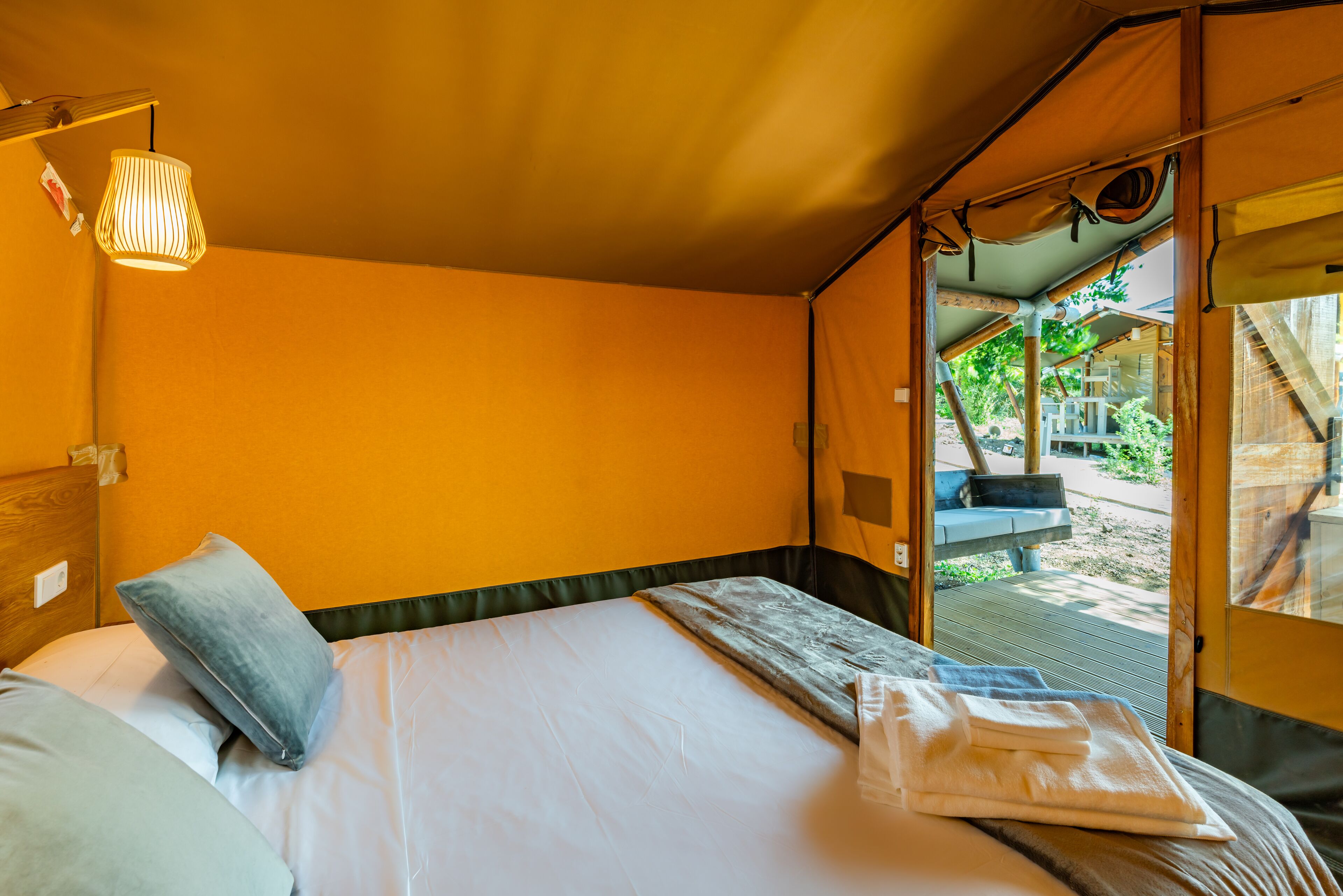 A single bed with a pillow, a blanket, and a folded towel. There is a window with a view of the outdoors.