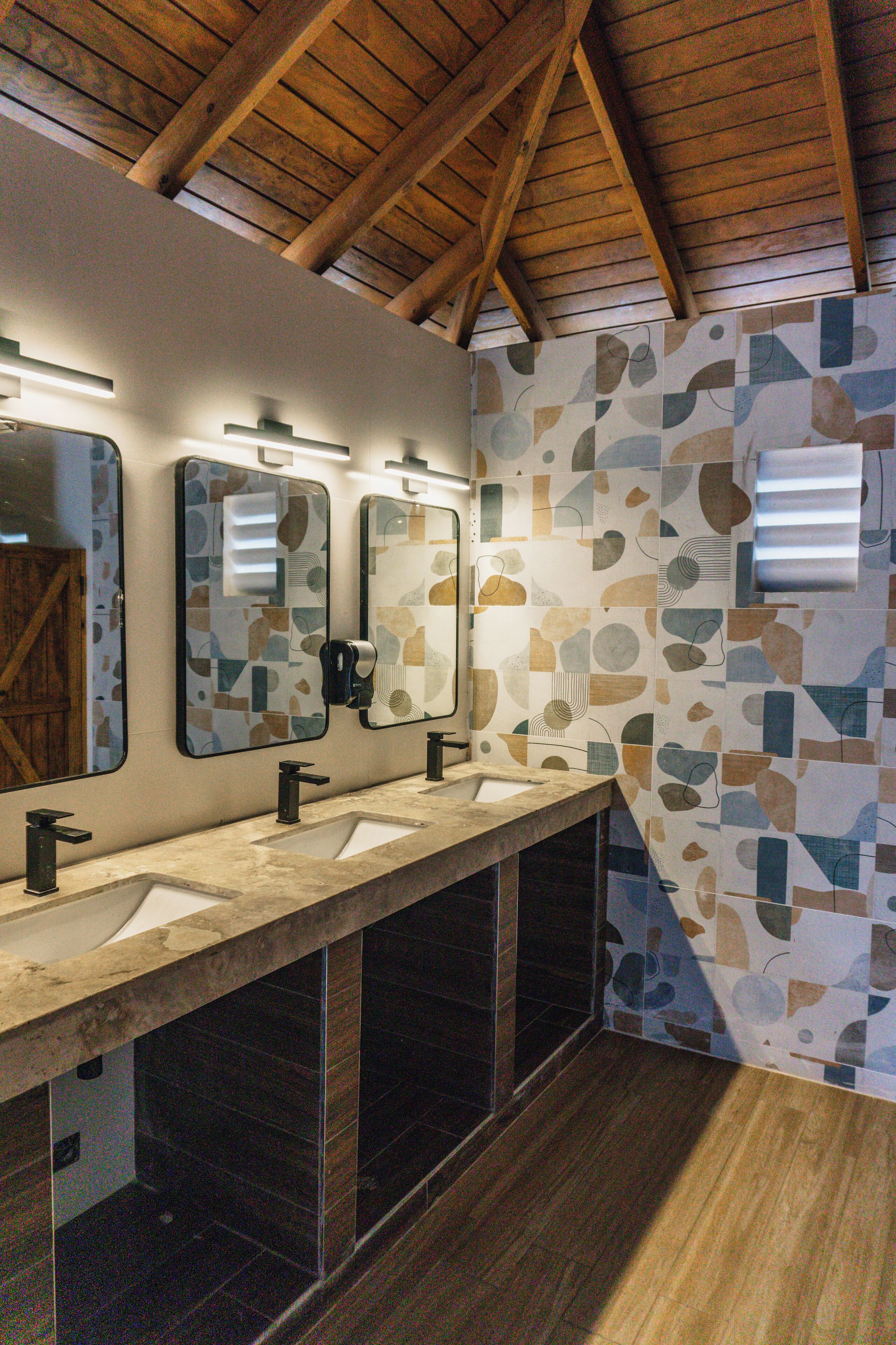 A bathroom with a long vanity, three sinks, and a mirror above each sink. The walls are tiled with a geometric pattern.