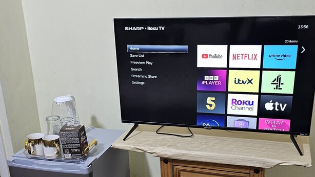 A television on a wooden stand with a menu interface, a small table with a kettle and cups, and a grey cabinet. - Beautiful 1-bed House in Leeds (Leeds)