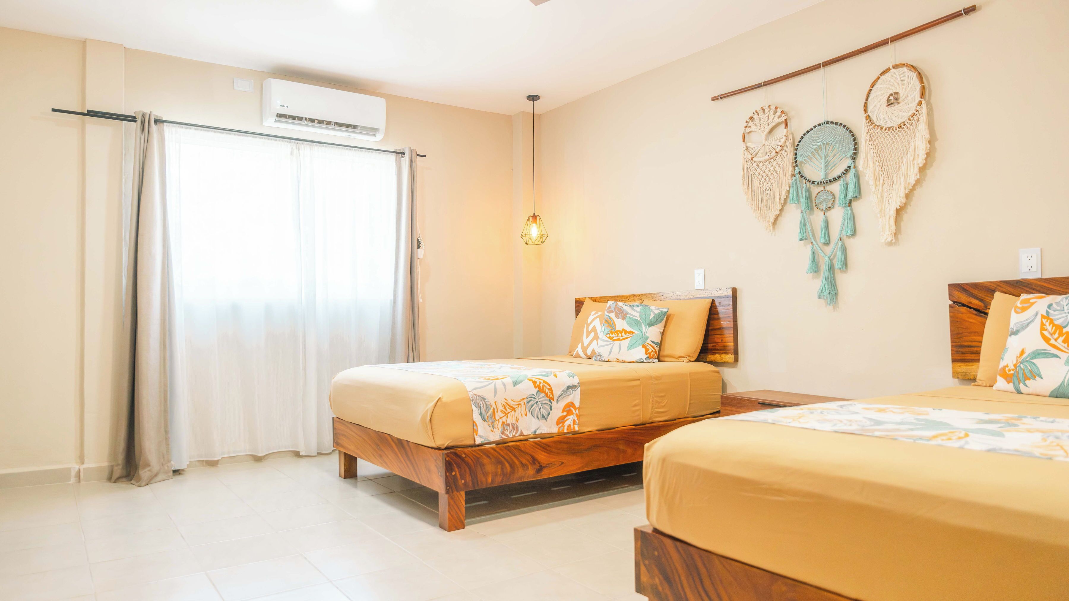 Ein Hotelzimmer mit zwei Betten, einem Deckenventilator, einer Klimaanlage, einer wandmontierten Lampe und dekorativen Wandbehängen aus Makramee.