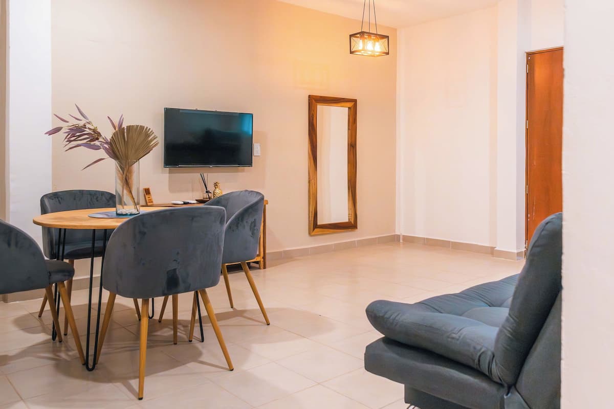 Een moderne woonkamer met een ronde eettafel, twee grijze fauteuils, een bank, een flatscreen televisie en een decoratieve vaas.