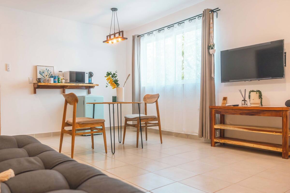 Een moderne woonkamer met een bank, een eettafel met stoelen, een televisie op een dressoir, en een keukenhoek met een magnetron en een plant.