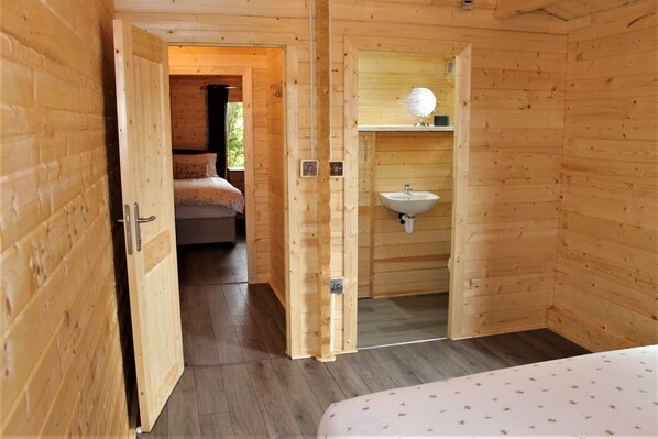 Habitación de cabaña de madera con una cama, una puerta de madera que da a otra habitación y un baño con lavamanos y espejo.