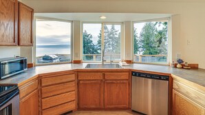 Una cocina con armarios de madera, lavavajillas de acero inoxidable y un ventanal con vistas a un balcón donde se ven árboles y una masa de agua.