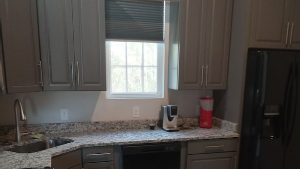 A kitchen with grey cabinets, a black refrigerator, a dishwasher, a coffee maker, and a window with blinds. - Beautiful retreat with gameroom and outdoor grill. (Myrtle Beach)