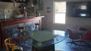 A dining area with a glass-top table, wooden barrel base, and two yellow chairs. A ceiling fan and a bar with stools are visible in the background. - Beautiful retreat with gameroom and outdoor grill. (Myrtle Beach)