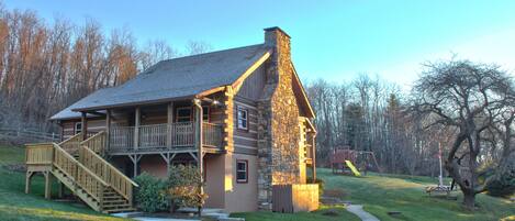 A log cabin with a stone chimney, a wooden deck, and a staircase leading to a balcony.