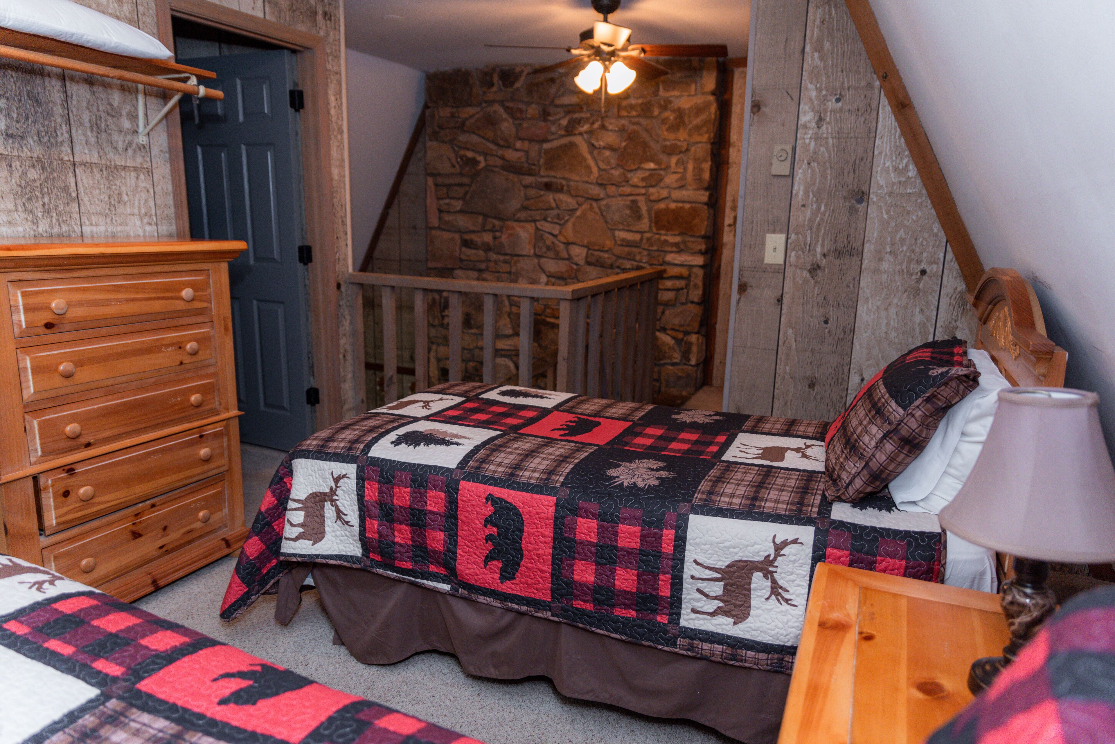 A bedroom with a wooden dresser, a bed with a quilt featuring animal prints, a stone wall, and a wooden staircase.