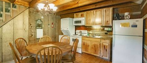 A wooden interior with a dining table and chairs, a kitchen with wooden cabinets, a white refrigerator, and a chandelier.