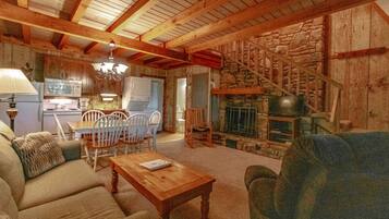 A cozy living room with a fireplace, wooden beams, a dining area with chairs, and a kitchen in the background.