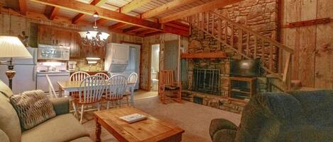 A cozy living room with a fireplace, wooden beams, a dining area with chairs, and a kitchen in the background.