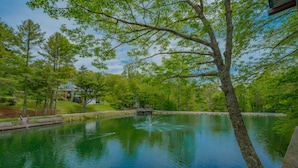 A serene lake with a fountain, surrounded by lush greenery and a wooden deck. - Swiss Mountain Village (Blowing Rock)
