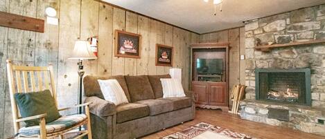 A cozy living room with a wooden wall, a stone fireplace, a wooden rocking chair, a sofa, and a patterned rug.