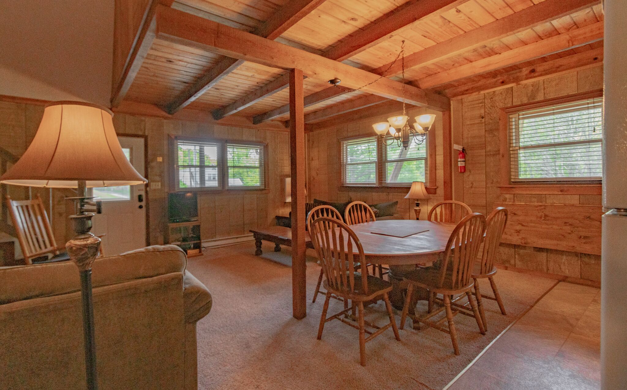 A cozy cabin interior with a wooden dining table surrounded by chairs, a sofa, and a lamp.
