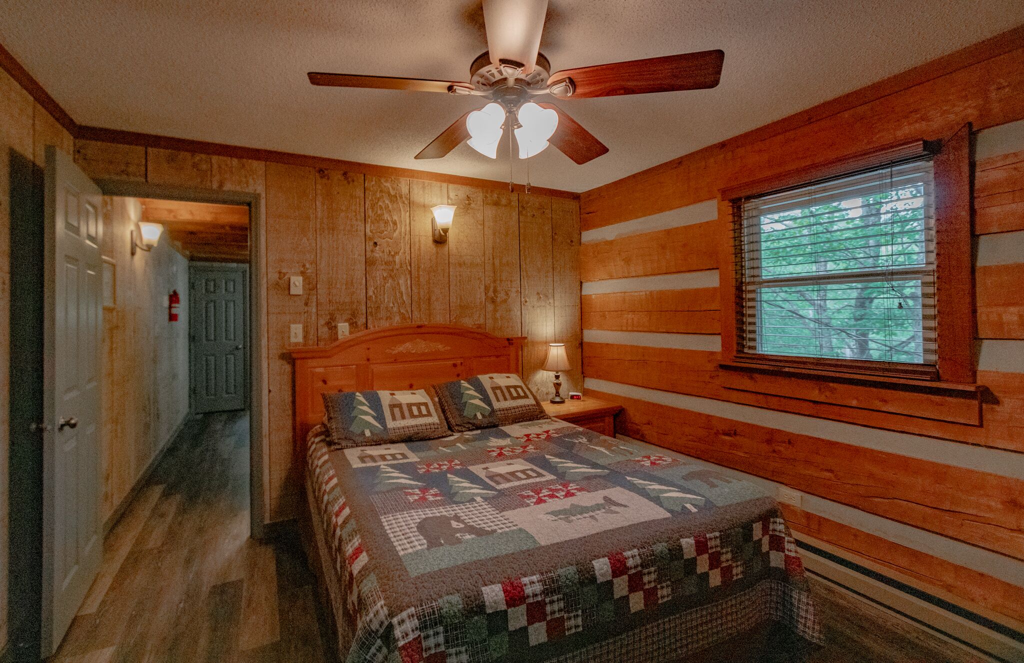 A bedroom with a wooden wall, a bed with a quilt, a ceiling fan, and a window with blinds.