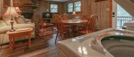 A cozy living room with a stone fireplace, wooden walls, a dining table with chairs, a sofa, a TV, and a hot tub.