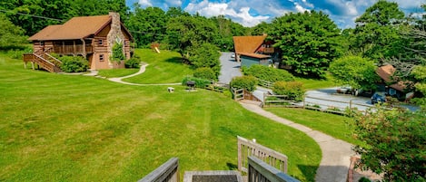 A rustic lodge with a stone chimney, a wooden deck, and a winding driveway surrounded by lush greenery.