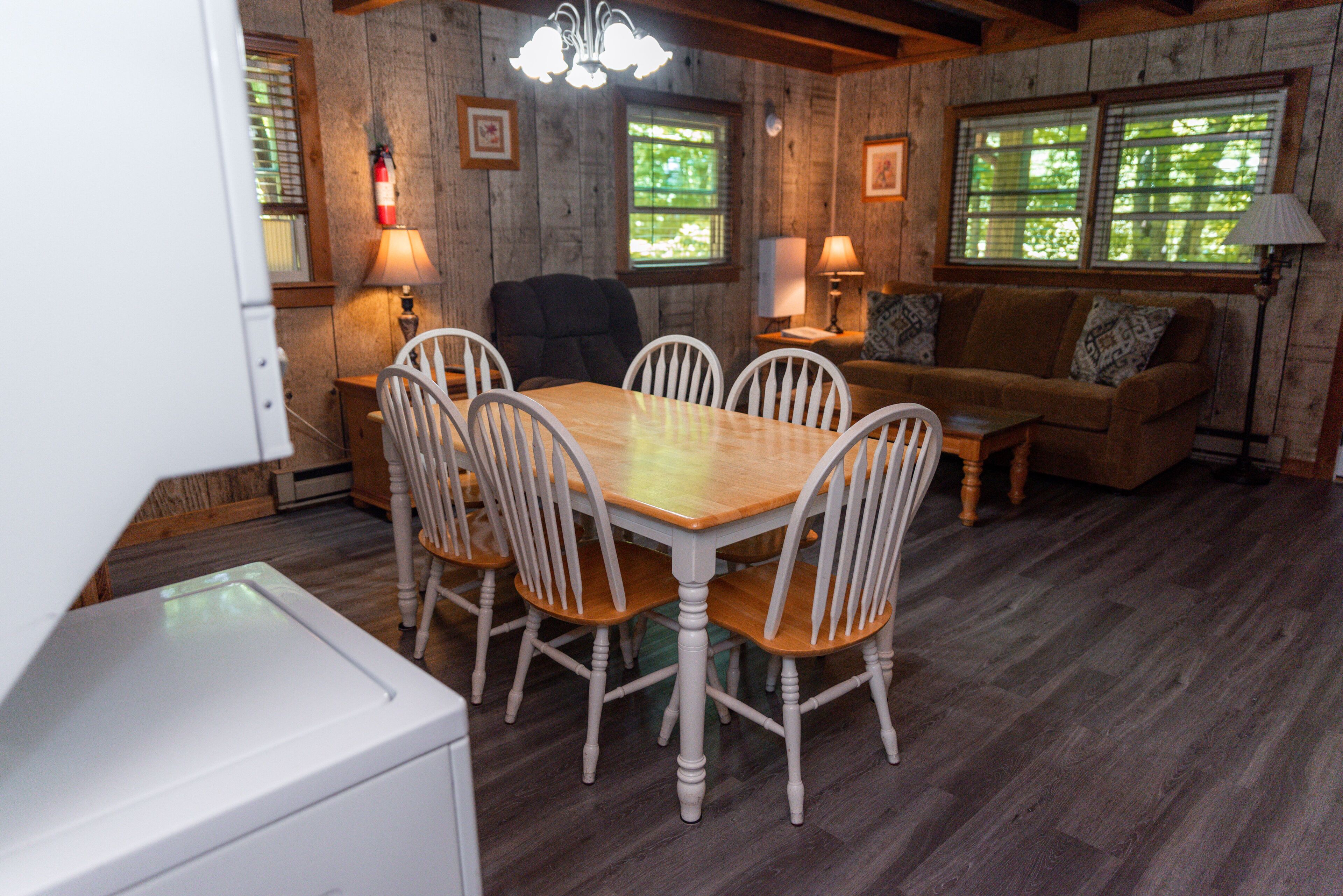 A dining area with a wooden table and white chairs, a sofa with patterned cushions, and a window with blinds.