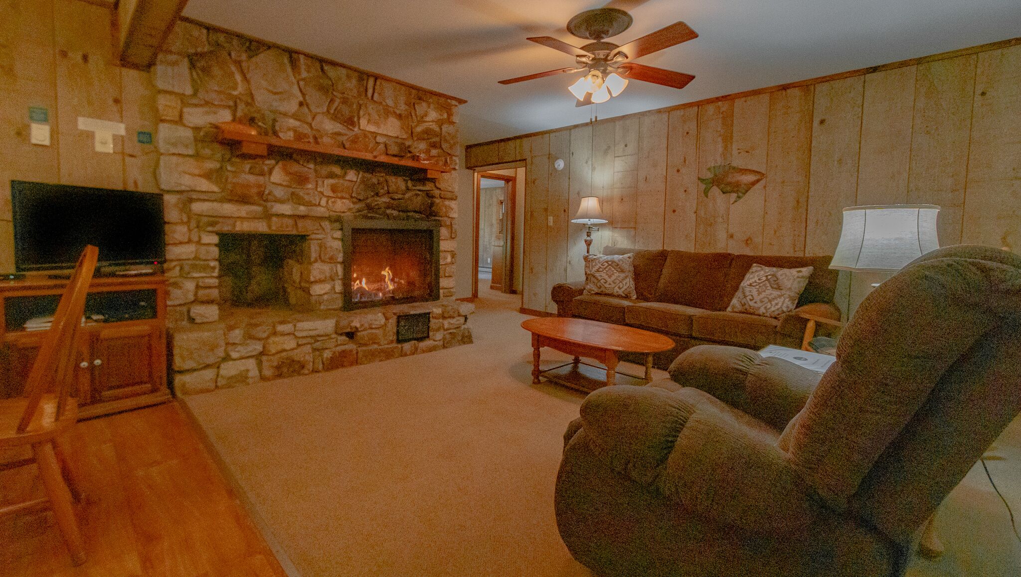 A cozy living room with a stone fireplace, a television, a sofa, and a ceiling fan.