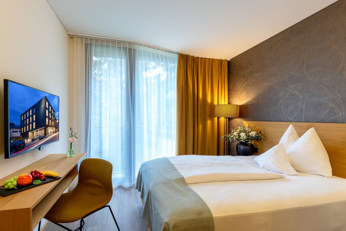 Ein Hotelzimmer mit einem Bett, einem Schreibtisch mit Stuhl, einem Fernseher und einem Fenster mit Vorhängen.