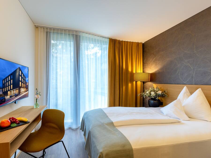 A hotel room with a bed, a desk with a chair, a television, and a window with curtains.