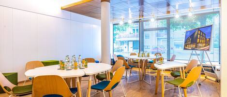 A modern dining area with round tables and chairs, a large window with blinds, and a projection screen displaying a building image.