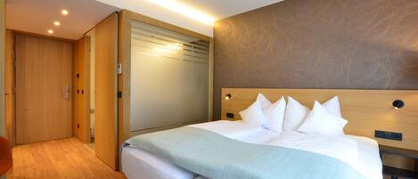 A modern hotel room with a large bed, wooden flooring, a built-in wardrobe, and a wall-mounted headboard.